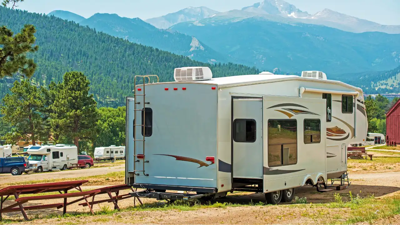 緑が広がる山を背景でキャンプ場にキャンプカーや乗用車をバックに白いキャンピングトレーラーとウッドテーブルが正面にある画像