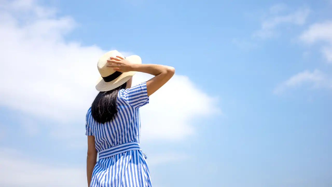 青空を見上げながら帽子に手を当てている女性の後姿の画像