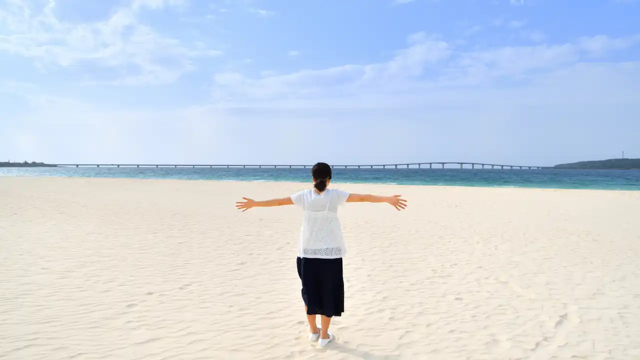 青空と海が広がる砂浜に女性が立って両手を広げ景色を感じている後ろ姿画像