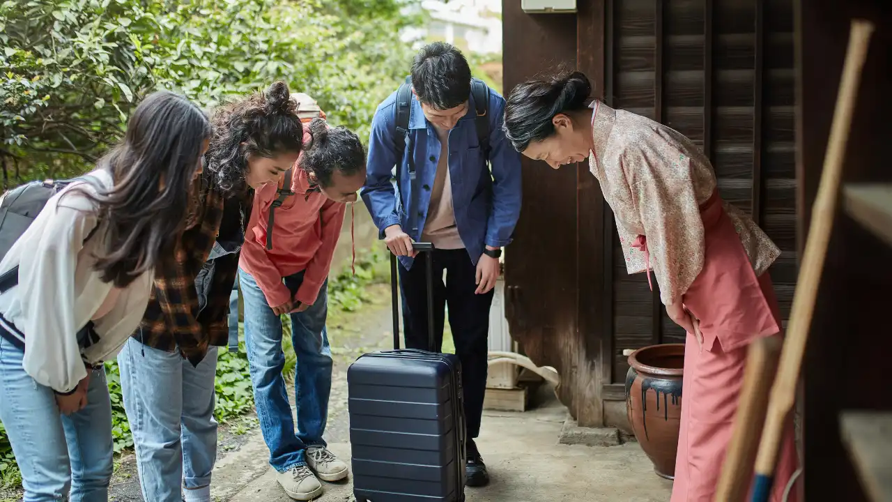 旅館の外。スーツケースを持った男性と大きなリュックを背負った3人の人々。旅行客を笑顔でお辞儀して出迎える旅館スタッフの画像